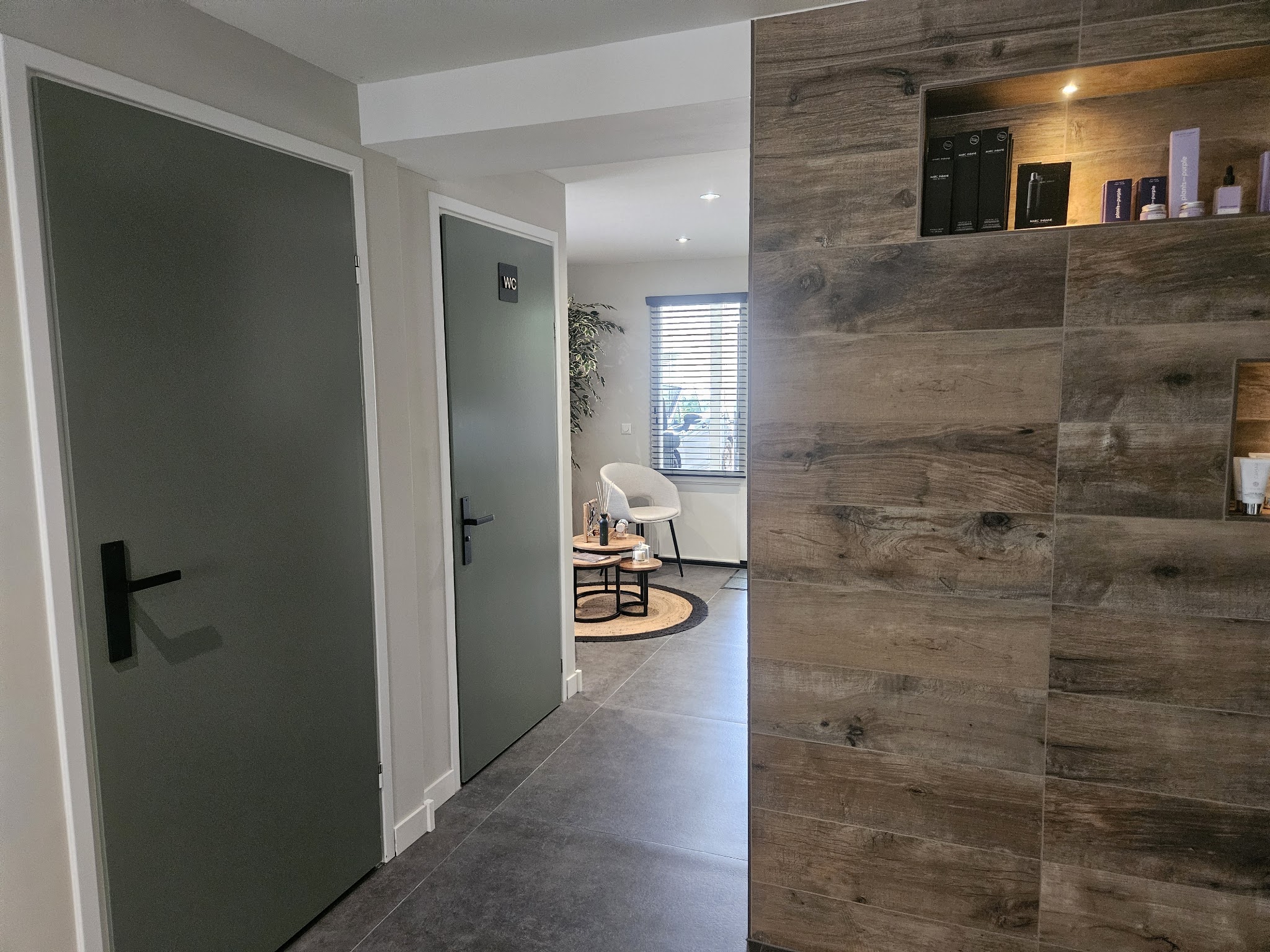 De gang met grijze deuren en een houten wand met een verlichte nis, achterin de zithoek